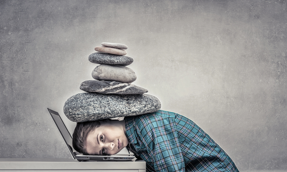 Frau sitzt vor Laptop, ihr Kopf liegt auf der Tastatur, sie hat einen Stapel Steine im Nacken.