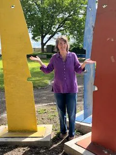 Heide in lila Bluse steht in einer Skulptur aus aus einem roten, einem gelben und einem hellblauen Kopf aus Metall. Die Köpfe sind zweidimensional, einander zugewandt, Heide steht zwischen dem gelben und dem roten Kopf.
