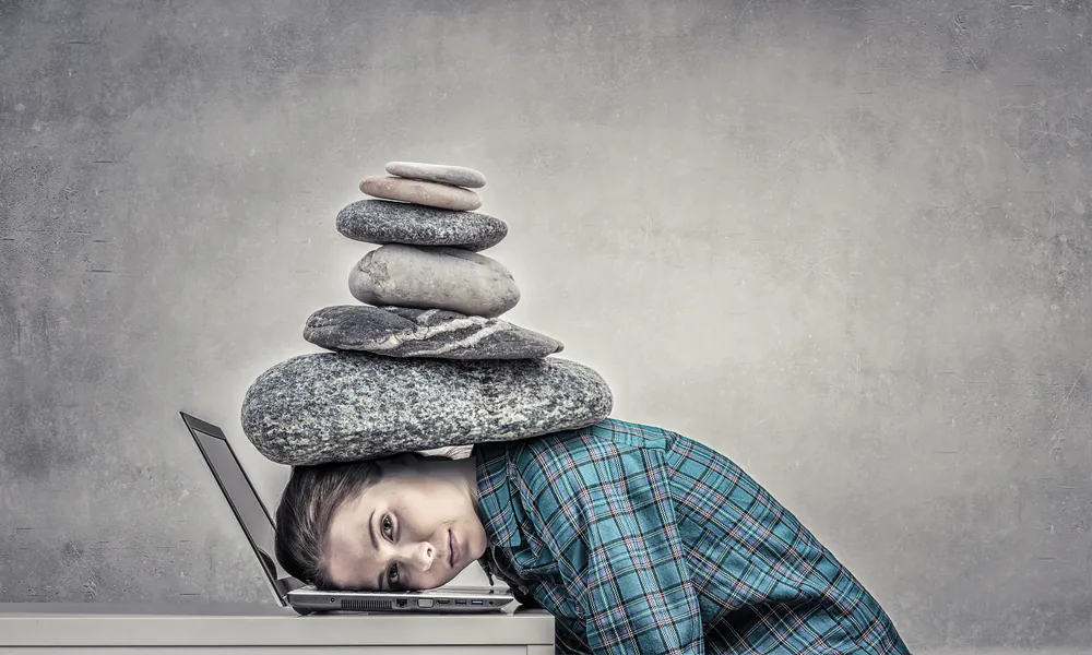 Frau sitzt vor Laptop, ihr Kopf liegt auf der Tastatur, sie hat einen Stapel Steine im Nacken.